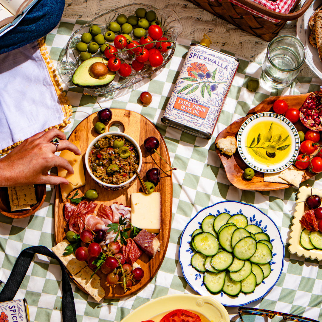 An overhead lifestyle shot of a picnic spread on a green and white checkered blanket, featuring charcuterie boards, sliced cucumbers, tomatoes, olives, and a vintage book titled 'Spicimarra'.