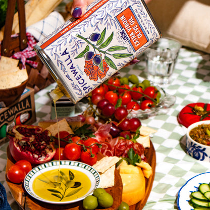 A hand holds a tin of Speckwalla extra virgin olive oil over a charcuterie board with tomatoes, cheese, olives, and bread on a checkered tablecloth.
