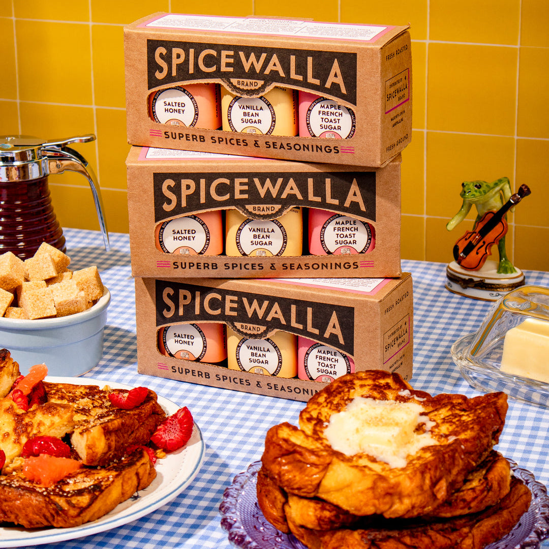 Three boxes of Spicewalla Superb Spices & Seasonings stacked on a table, surrounded by breakfast foods like French toast, toast with butter, and cubed bread, with a yellow tiled wall background.