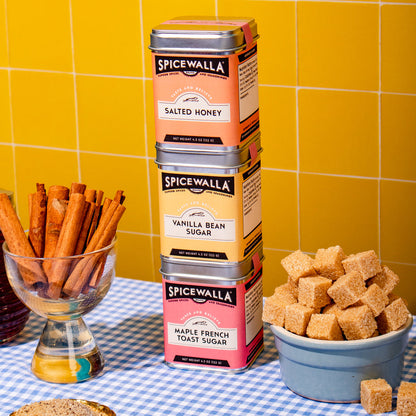 Four stacked Spicywalla tea tins on a blue-and-white checkered tablecloth in front of a yellow tiled wall. Surrounding props include a glass bowl of cinnamon sticks, a bowl of sugar cubes, and a small dish of loose sugar.