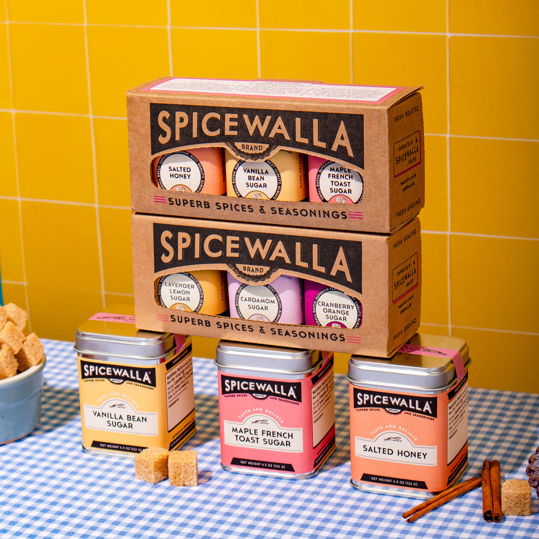 A styled product display of Spicewalla Super Spices & Seasonings, featuring two large brown cardboard boxes stacked on top of three matching metal tins on a blue-and-white checkered tablecloth, set against a bright yellow tiled wall.