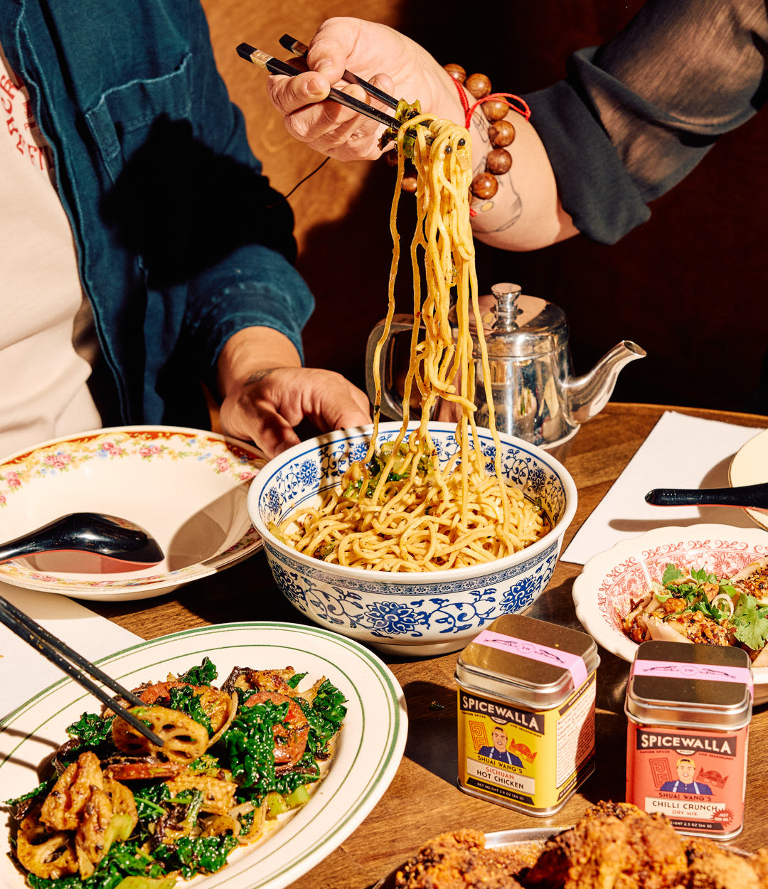 Two people sharing a bowl of noodles at a table with various dishes and condiments.