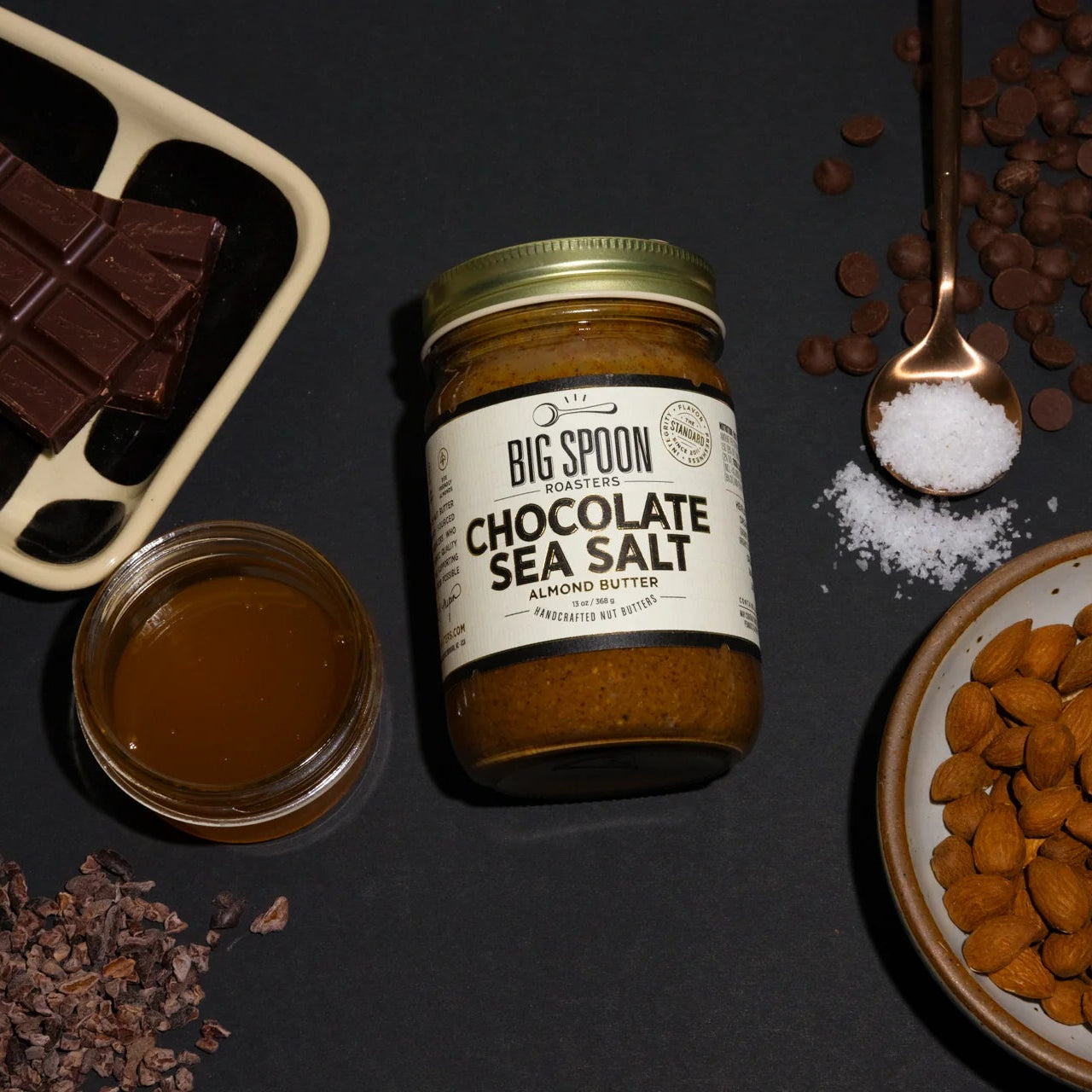 Jar of 'Big Spoon' Chocolate Sea Salt on a dark surface with chocolate pieces and sea salt.