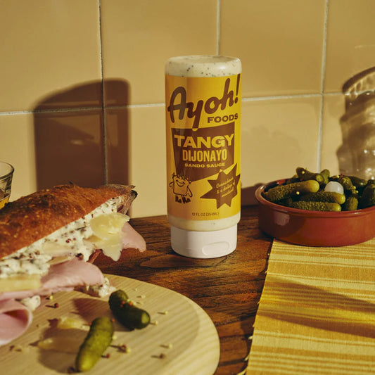 Ayoh! Foods Tangy Dijonayo condiment bottle with a sandwich and pickles on a wooden surface.