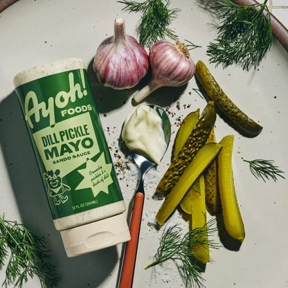 Ayoh! Foods Dill Pickle Mayo on a plate with pickles, garlic, and herbs.