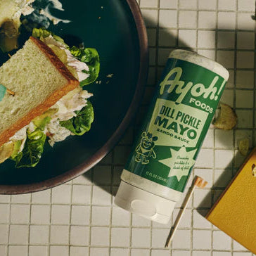 Ayoh! Dill Pickle Mayo container with a sandwich and chips on a checkered tablecloth.