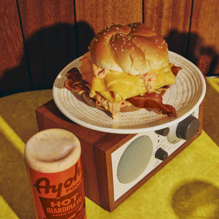 Hamburger on a plate with a small radio and Ayala hot sauce bottle on a table.