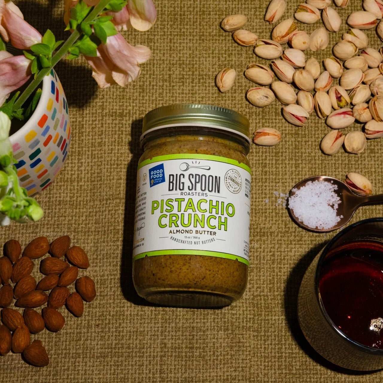 Jar of Big Spoon Pistachio Crunch Almond Butter surrounded by pistachios and almonds on a textured surface.