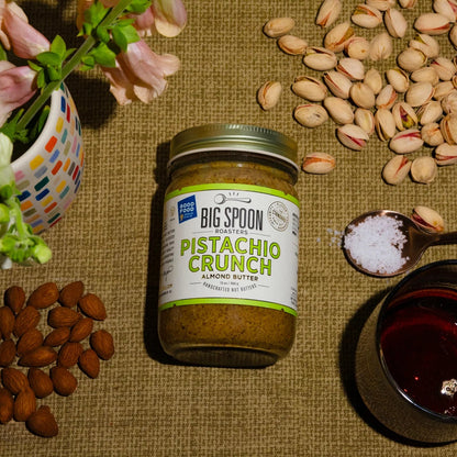 Jar of Big Spoon Pistachio Crunch Almond Butter surrounded by pistachios and almonds on a textured surface.