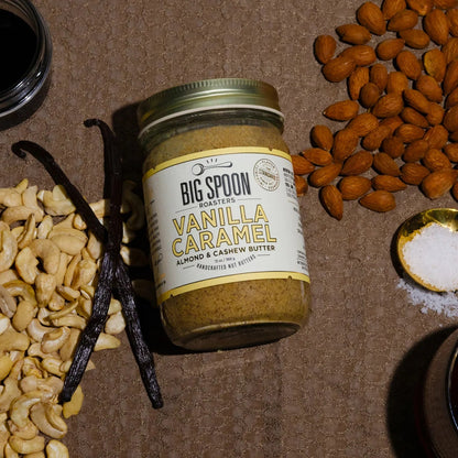 Jar of Big Spoon Vanilla Caramel Nut Butter surrounded by cashews, vanilla beans, and almonds on a brown surface.