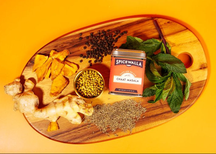 Spices and herbs on a wooden board with a can of SpiceWala Chat Masala against an orange background